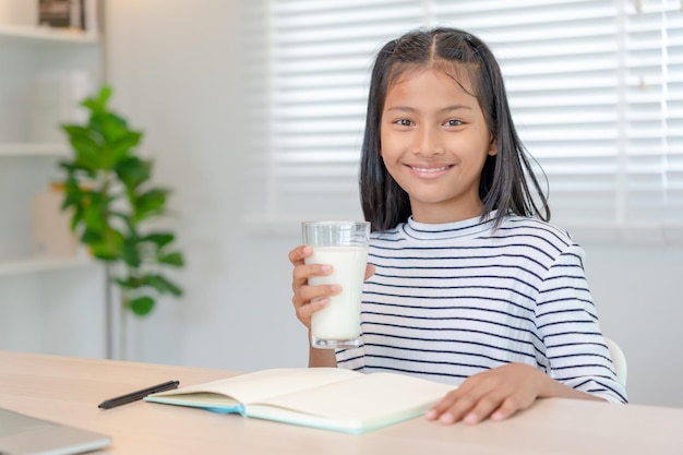 Kinder Frauen trinken Milch, um den Körper zu nähren und das Gehirn zu nähren. Asiatisches junges kleines Mädchen lernt zu Hause, Mädchen trinkt glücklich Milch und liest ein Buch für die Prüfung. Homeschool-Erziehung, Vitaminentwicklung