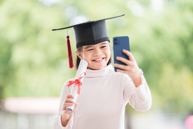 Kinder graduieren mit Abschlusskappe und gerolltem Abschlusszertifikat mit Smartphone