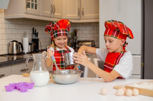 Kinder in Kochkostümen bereiten Teig aus Mehl, Milch, Eiern und Butter für Weihnachtsgebäck zu. bruder und schwester kochen festliches abendessen, kekse in der küche. Winterkonzept des neuen Jahres