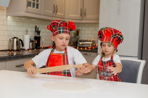 Kinder in Kochkostümen rollen den Teig mit einem Nudelholz für Weihnachtsplätzchen aus