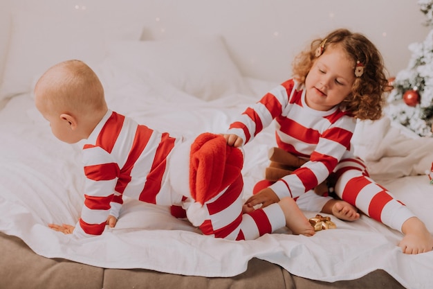 Kinder in rot-weißen Schlafanzügen spielen, haben Spaß, schneiden Grimassen im Bett. bruder und schwester, junge und mädchen, die weihnachten feiern. Lebensstil. Platz für Text. Foto in hoher Qualität