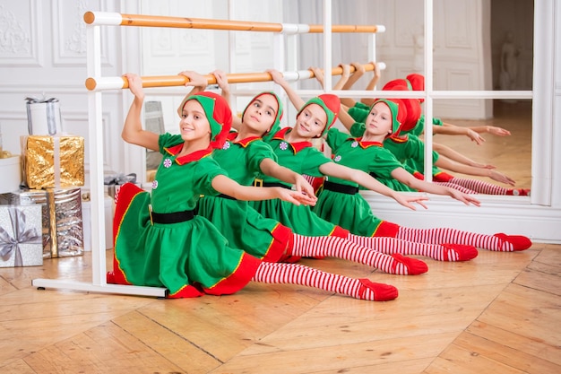 Kinder in Weihnachtskostümen von Weihnachtsmann-Elfen trainieren an der Ballettstange in einem geräumigen weißen Studio.