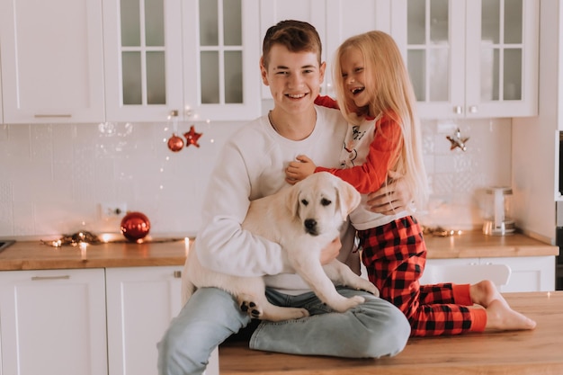 Kinder mit ihrem geliebten Hund sitzen in der Weihnachtsküche. Familienzeitvertreib. Lebensstil. Bruder und Schwester, Junge und Mädchen spielen und albern mit einem Haustier herum. Platz für Text. Foto in hoher Qualität