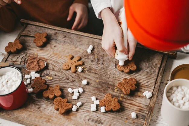 Kinder schmücken den Weihnachtslebkuchenmann mit weißem Zuckerguss auf einem hölzernen Hintergrund. Weihnachtssüßigkeiten mit Kakao und Marshmallows für Kinder. Winterurlaub zu Hause. Spaß in der Küche.