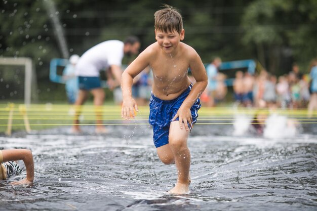 Kinder spielen mit Wasser, Junge schwimmt in Seifenblasen, Sommergarten, Freiluft für Kinder, Junge spritzt Wasser an einem heißen, sonnigen Tag.