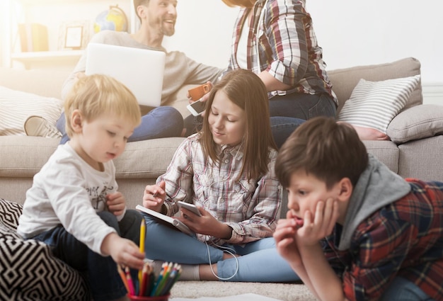 Kinder spielen und benutzen Geräte auf dem Boden, während sich die Eltern mit Laptop auf dem Sofa entspannen