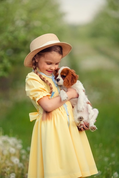 Kinder und Haustiere süßes kleines Mädchen in einem Hut mit Zöpfen, das einen braunen Welpen Cavalier King Charles Spaniel in ihren Armen hält