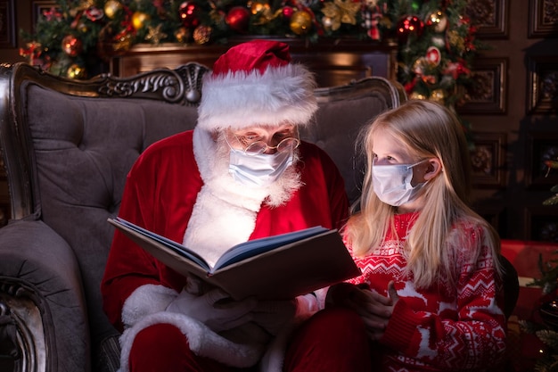 Kinder und Weihnachtsmann in medizinischen Masken Mädchen und Weihnachtsmann lesen Weihnachtsbuch in der Nähe des Weihnachtsbaums zu Hause Frohes neues Jahr Covid 2021