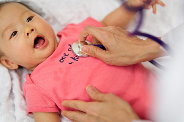 Kinderarzt hört Herzfrequenz des netten neugeborenen Babys, das auf dem Bett unter Verwendung des Stethoskops liegt.