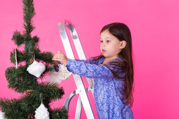 Kinderferien und Weihnachtskonzept kleines Mädchen, das Weihnachtsbaum auf rosa Hintergrund verziert