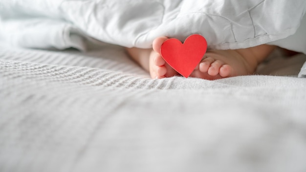 Kinderfersen, Baby, liegen auf dem Bett unter der Decke mit einem Etikett am Finger, mit einem Herz. Valentinstag.