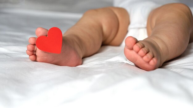 Kinderfersen mit einem Etikett am Finger, mit Herz, liegen auf dem Bett auf weißem Hintergrund. Valentinstag.