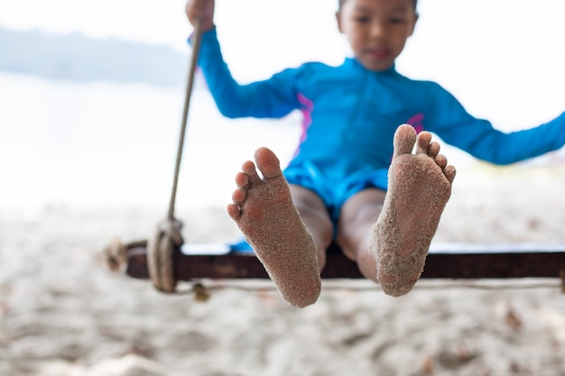 Kinderfüße mit Sand, während sie auf einer Schaukel am Strand nahe dem Meer in den Sommerferien spielt