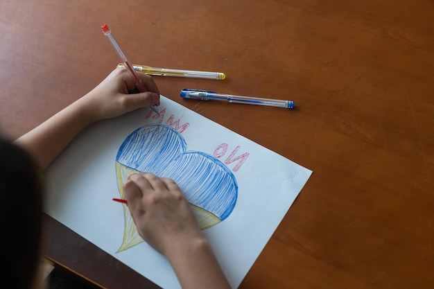 Kinderhände eines Kindes zeichnen eine herzförmige Ikone mit dem Bild der Nationalflagge der Ukraine. Von oben betrachten. Kinder gegen Krieg. Kinderzeichnung für den Frieden in der Ukraine