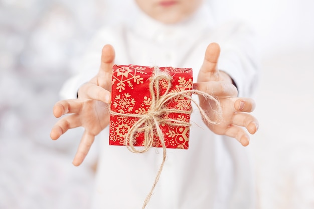 Kinderhände halten Geschenkbox eingewickelt in Kraft rotes Papier gebunden. Urlaubskonzept. Weihnachtsferien.