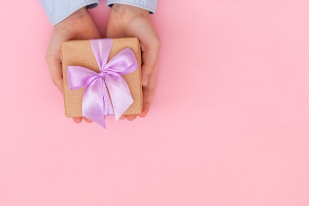 Kinderhände halten Geschenkbox in Bastelpapier gewickelt und mit Schleife an rosa Wand gebunden.