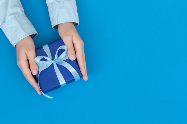 Kinderhände halten Geschenkbox in Bastelpapier gewickelt und mit Schleife auf blauem Konzept Vatertag oder Geburtstagsgrußkarte gebunden