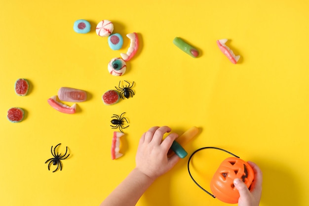 Kinderhände sammeln Marmeladen für Halloween mit einem kürbisförmigen Korb auf gelbem Hintergrund mit Platz für Text