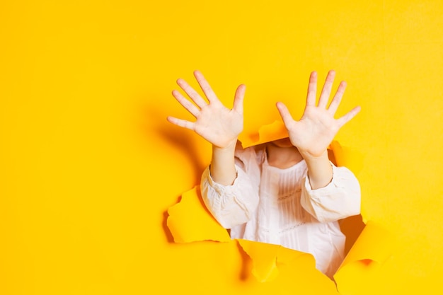 Kinderhände zeigen zehn Finger durch ein zerrissenes Loch in gelbem Papier mit Kopierraum Gesten und Körpersprache