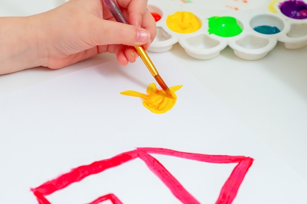 Kinderhand, die gelbe Sonne mit rotem Haus durch Aquarelle auf weißem Blatt Papierhand des Kindes zeichnet