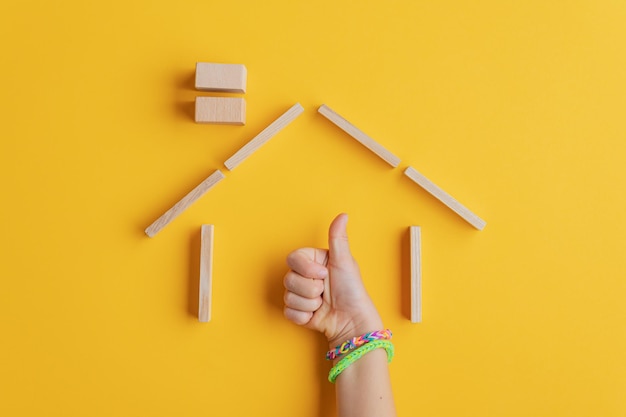 Kinderhand mit bunten Armbändern, die ein Daumen-hoch-Zeichen machen, das seine Hand in einem Haus aus Holzpflöcken hält. Über gelbem Hintergrund.