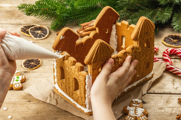 Kinderhand schmücken das Lebkuchenhaus mit süßem Zuckerguss. Handgefertigter Prozess, festliches Neujahrsdekor. Traditionelle Gewürze, Holzhintergrund, Nahaufnahme