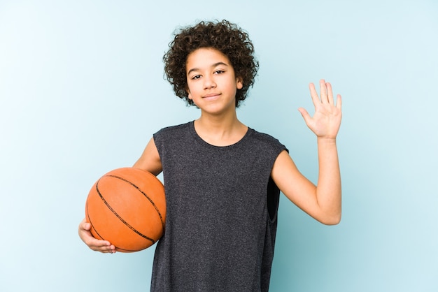 Kinderjunge, der Basketball spielt, isoliert auf der blauen Wand lächelnd fröhlich zeigt Nummer fünf mit den Fingern.