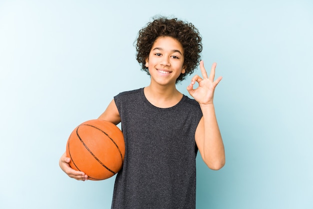 Kinderjunge, der Basketball spielt, lokalisiert auf blauer Wand fröhlich und zuversichtlich zeigt ok Geste.