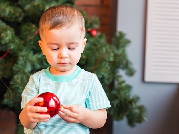 Kinderjunge, der eine rote Weihnachtsdekoration gegen Weihnachtsbaum hält