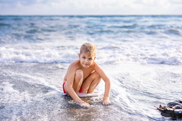 Kinderjunge, der in den Wellen auf dem Strand im Sommersonnenuntergang spielt, aufpassende Meereswellen des Kindes und Spaß hat