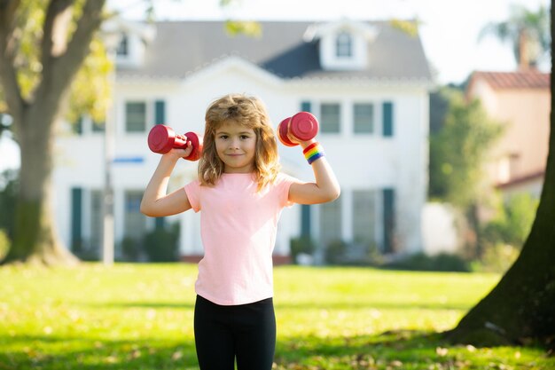 Kinderjunge, der mit Hanteln auf Parkhintergrund trainiert Kindersport im Freien lustiger aufgeregter Kindersport