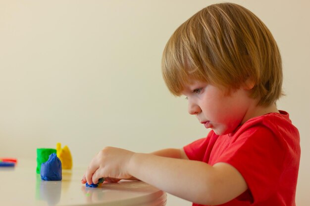 Kinderjunge, der mit Plastilin spielt, während er am Tisch sitzt, Seitenansicht, Kindheitsentwicklungsaktivitäten für Kinder