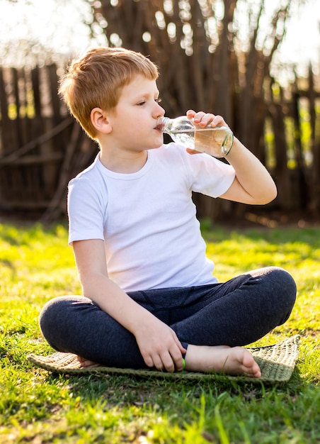 Kinderjunge im weißen T-Shirt, das sauberes kaltes Wasser aus der Flasche trinkt, die auf grünem Gras in der Natur sitzt