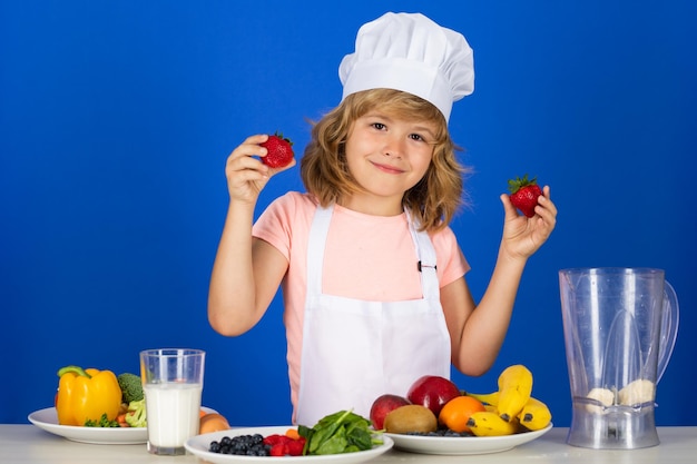 Kinderjunge in Kochmütze und Schürze halten Erdbeeren, die kochen, Mahlzeit zubereiten kleiner Koch mit Gemüse a