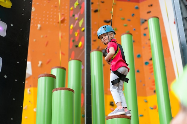 Kinderklettern an der Wand im Vergnügungszentrum Klettertraining für Kinder Kleines Mädchen in gekleideter Kletterausrüstung klettert hoch Extreme aktive Freizeit für Kinder