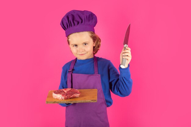 Kinderkoch hält Schneidebrett mit Fleisch, Rindersteak und Messer. Junger Koch, der gesunde Lebensmittel zubereitet. Porträt eines kleinen Kindes mit Kochmütze, isoliert auf Studiohintergrund. Kinderkoch. Kochprozess