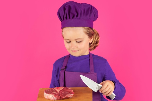 Kinderkoch hält Schneidebrett mit Fleisch, Rindersteak und Messer Kind trägt Herduniform und Kochmütze, die Essen im Studioporträt zubereitet