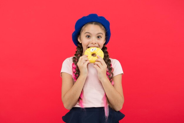 Kinderlächelndes Mädchen bereit, Donut zu essen. Süßwarenladen und Bäckereikonzept. Kinder sind große Fans von gebackenen Donuts. Unmöglich, frisch zubereiteten Donuts zu widerstehen. Mädchen halten glasierten süßen Donut in der Hand auf rotem Hintergrund.