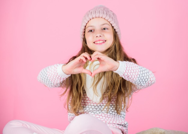 Kinderlanges Haar lächelnde Showherzgeste. Mädchen tragen Strickmütze rosa Hintergrund. Kümmere dich diesen Winter um Kopfhaut und Haare. Haarschäden im Winter vorbeugen. Tipps zur Haarpflege im Winter, die Sie befolgen sollten.