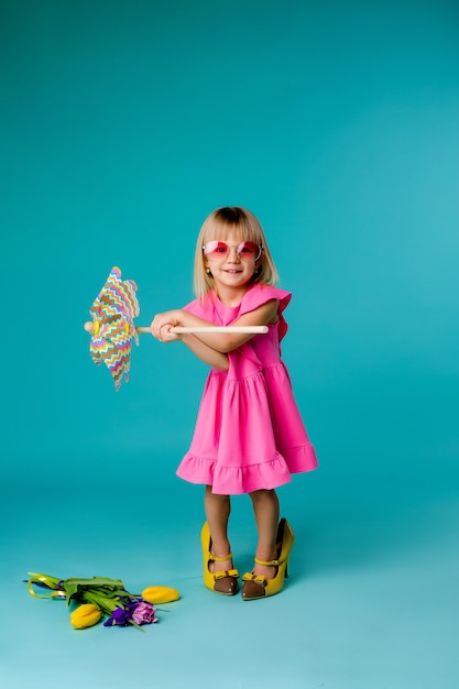 Kindermädchen, das in der Sonnenbrille lächelt, die eine Windmühle auf einem blauen Raum hält