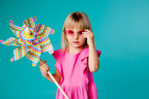 Kindermädchen, das in der Sonnenbrille lächelt, die eine Windmühle auf einem blauen Raum hält