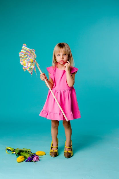 Kindermädchen, das in der Sonnenbrille lächelt, die eine Windmühle auf einem blauen Raum hält