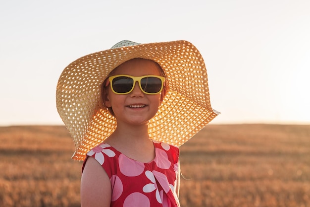 Kindermädchen im Strohhutkleid im Weizenfeld Lächelndes Kind mit Sonnenbrille bei Sonnenuntergang auf dem Land Ästhetik im Cottagecore-Stil