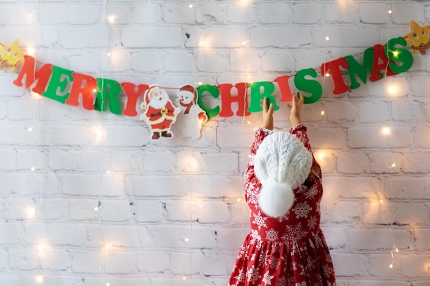 Kindermädchen in Strickmütze mit Pompon verzierte weiße Wand zu Hause Girlande Frohe Weihnachten