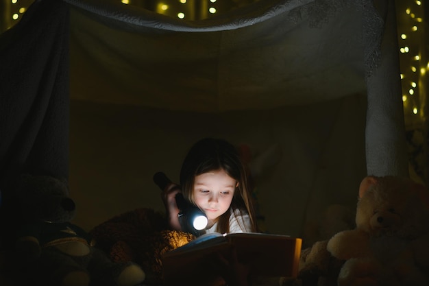 Kindermädchen lesen mit Buch und Taschenlampe und Teddybär im Zelt, bevor sie ins Bett gehen