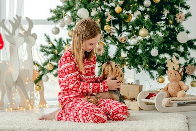 Kindermädchen mit dem Hund, der auf dem Boden mit Weihnachtsbaum im Hintergrund sitzt und Hündchen streichelt. Kind und Haustier genießen Neujahrszeit und Dekoration zu Hause