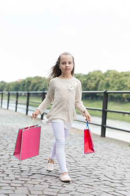 Kindermädchen mit langen Haaren, die gerne einkaufen. Fashionista-Mädchen, das mit rosa Taschen einkauft. Mädchen kauft gerne Kleidung. Einkaufskonzept. Mädchen, das auf ruhigem Gesicht einkauft, trägt Einkaufstaschen, städtischen Hintergrund