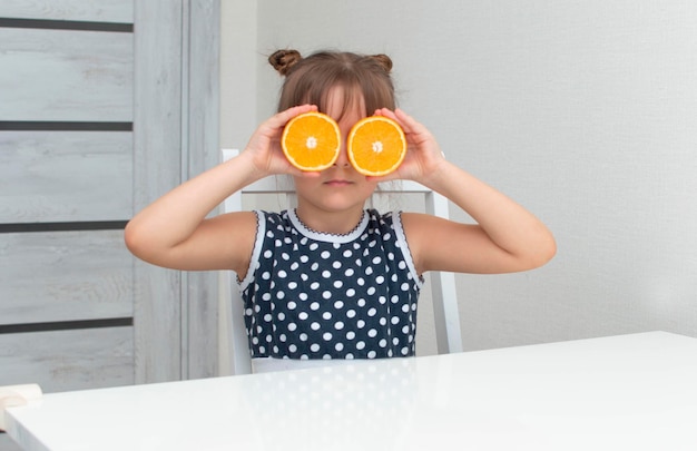 Kindermädchen mit Orangen auf den Augen, die am leeren Raumtisch mit einem Glas Orangensaft sitzen. Gesunde Vitaminkinderernährung. Kind mit Früchten. Lustiges Kleinkind.