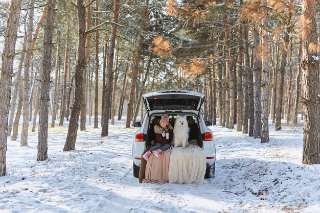 Kindermädchen sitzt im Kofferraum des Autos mit ihrem Haustier, einem weißen Hund Samojeden, im Winter im verschneiten Kiefernwald, ein Mädchen, das Tee von einer Thermoskanne trinkt