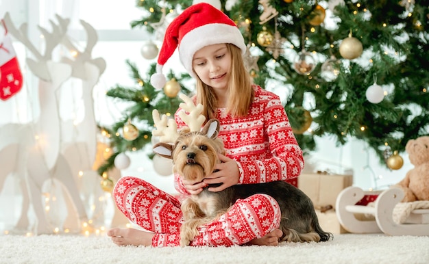 Kindermädchen und -hund mit Weihnachtsbaum im Hintergrund zu Hause. Kind trägt auf Hündchen-Haustier verzierten Hirsch-Neujahrsohren